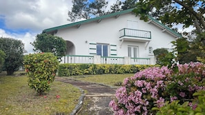 A white building with a green roof, a balcony, and a well-maintained garden with pink flowers.