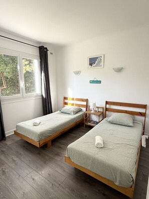 A room with two single beds, a small table with a lamp, a window with curtains, and a wall-mounted light fixture.