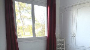 Ein ordentlich bezogenes Bett mit weißen Leintüchern, ein Fenster mit roten Vorhängen und ein weißer Kleiderschrank.
