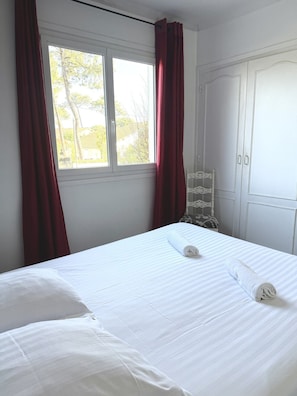Una cama bien hecha con ropa blanca, una ventana con cortinas rojas y un armario blanco.