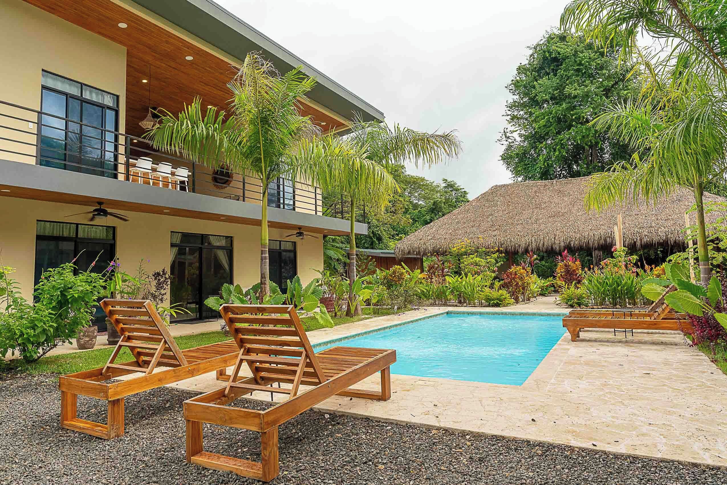 A modern house with a pool, wooden lounge chairs, and a thatched roof structure.