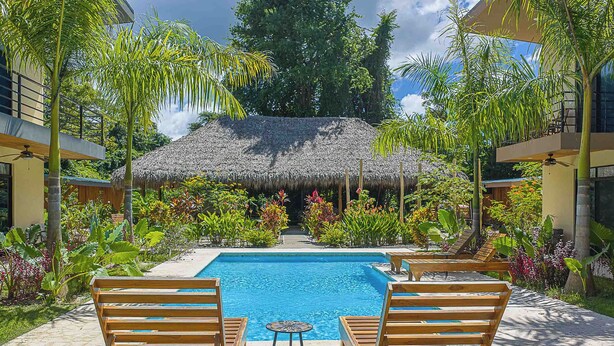 Ein Pool mit Holzstühlen, einem strohgedeckten Gebäude und tropischen Pflanzen.