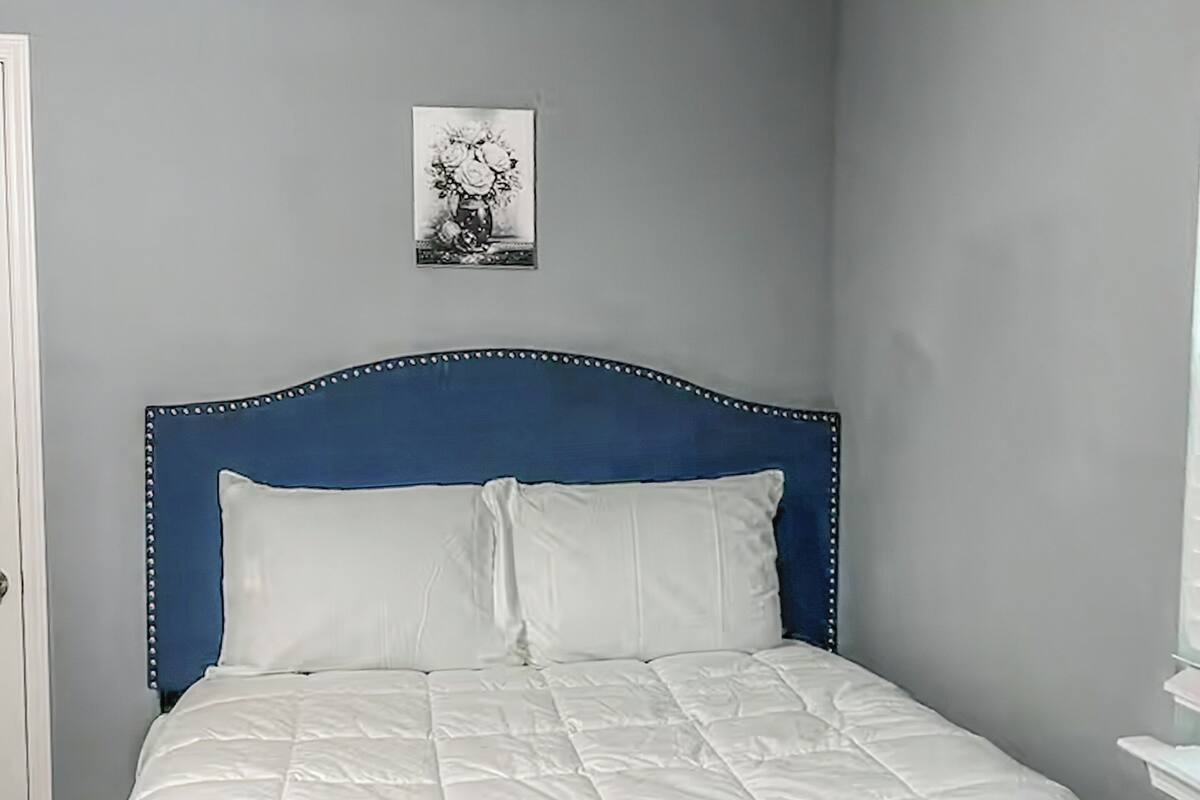 Una cama con cabecera azul, ropa de cama blanca y un cuadro en la pared.