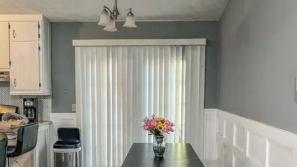 Uma sala de jantar com mesa e bancos, uma cozinha ao fundo e uma janela com persianas.
