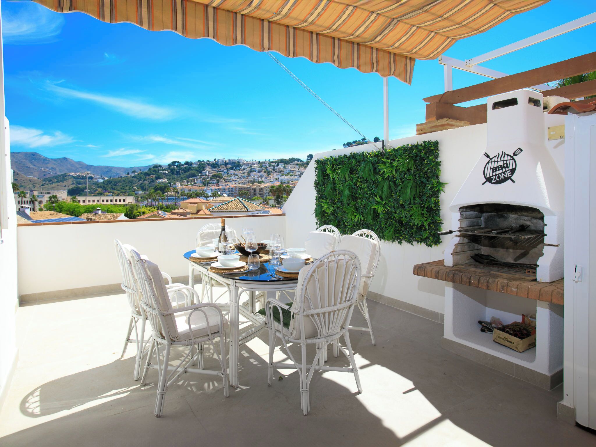 A patio with a table set for four, white wicker chairs, a BBQ grill, and a view of a cityscape.