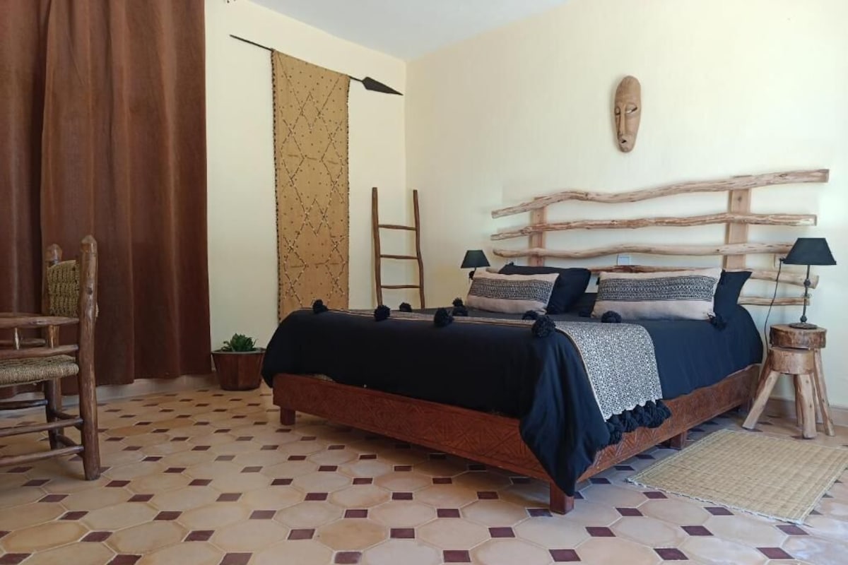 Een slaapkamer met een bed, een houten hoofdeinde, een ladder, een kamerplant en een gordijn.