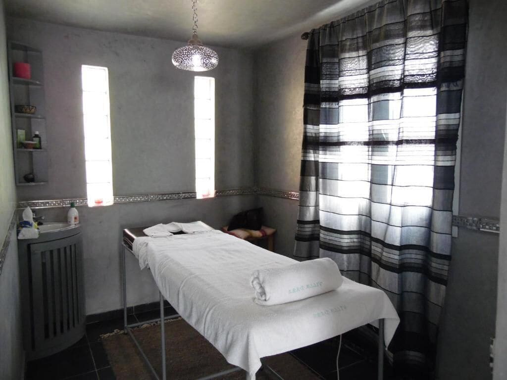 Quarto com uma cama de solteiro, radiador, prateleira com artigos de higiene e janela com cortinas.