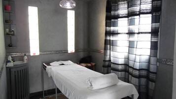 Quarto com uma cama de solteiro, radiador, prateleira com artigos de higiene e janela com cortinas.