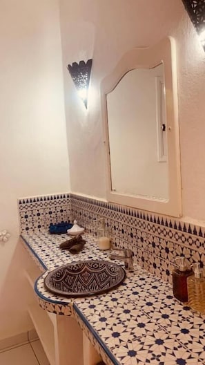 Een badkamer met een betegelde spatwand, een rond wastafel met decoratief bord, een spiegel en een wandlamp.