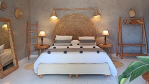 A bedroom with a large bed, two bedside tables with lamps, a wooden ladder, a woven headboard, and a wooden shelf with hanging items. - Villa Daba by Zelia en Exclusivité  (Essaouira)