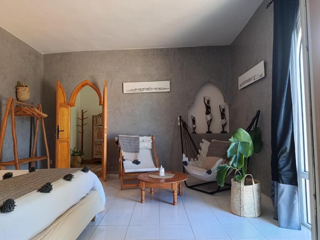 Une pièce comprenant un lit, une chaise en bois, une petite table, un fauteuil suspendu et une grande plante.