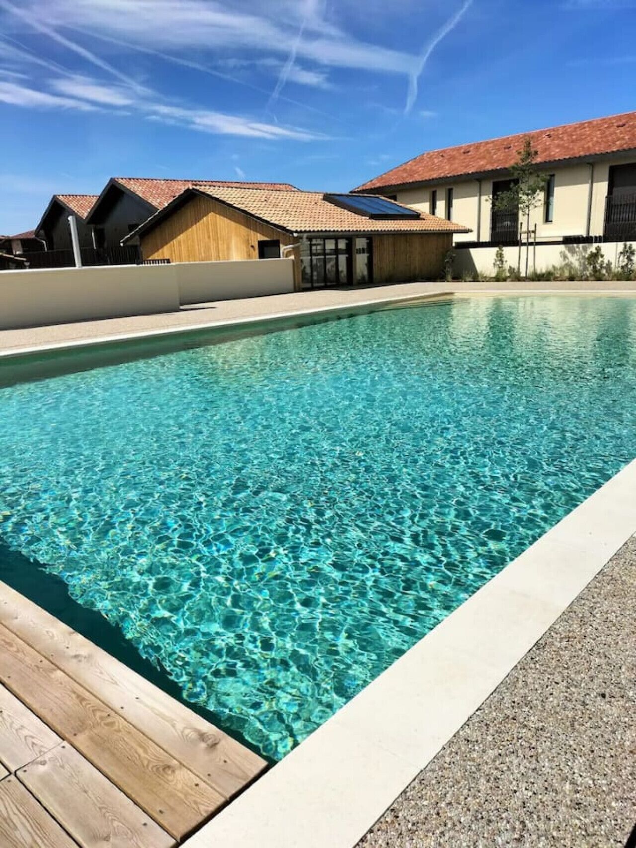 Ein klares Schwimmbecken mit einer Holzterrasse und einem Gebäude mit einem geflieselten Dach im Hintergrund.