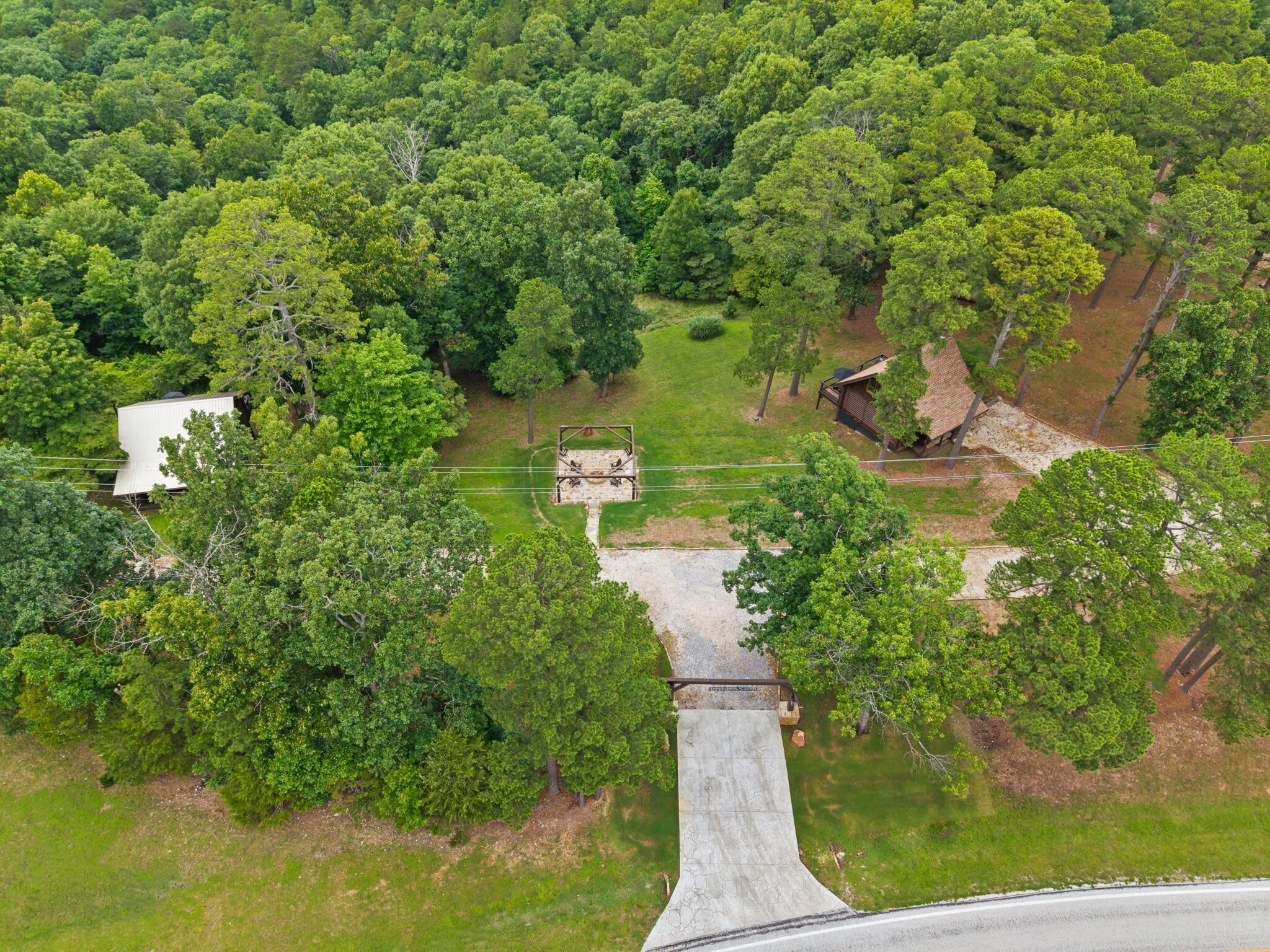 An aerial view of a property surrounded by dense forest, featuring a paved driveway leading to a building with a sloped roof.