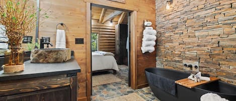 A log cabin-style bathroom with a stone wall, a black bathtub, and a wooden vanity.
