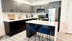 A modern kitchen with grey cabinets, a white countertop, a stainless steel refrigerator, a microwave, and a sink. There are two blue bar stools at the island. - Cozy Chic 1 Bdrm King Suite - mins to NYC, MetLife (Belleville)