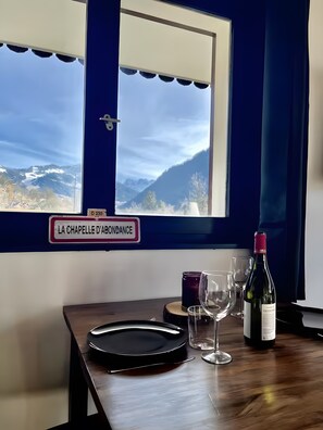 Ein Tisch mit einer Weinflasche, Gläsern und einem Schild mit der Aufschrift 'LA CHAPELLE D'ABONDANCE' in einem Raum mit Blick auf eine malerische Berglandschaft.