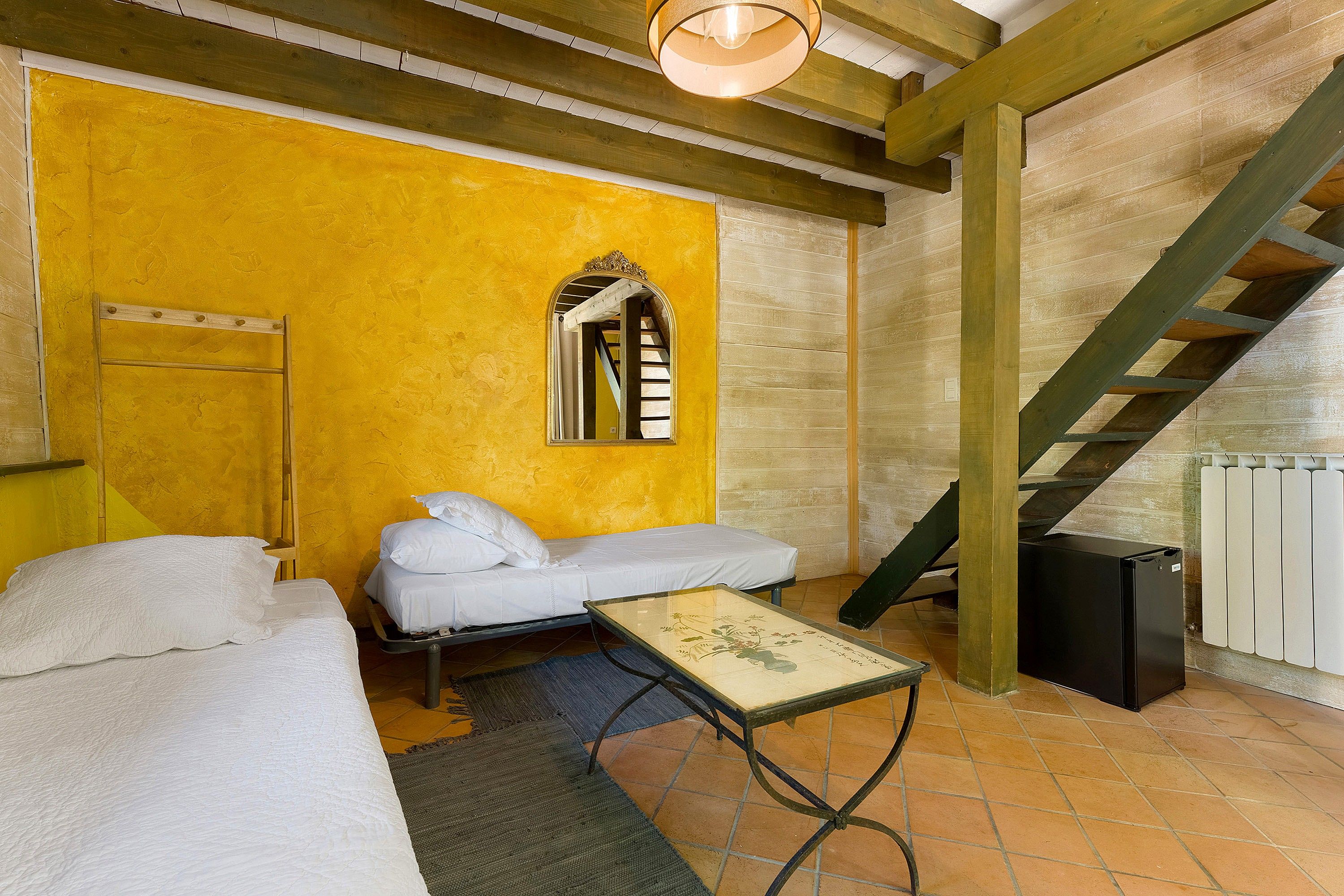 Ein Zimmer mit gelber Wand, einem Bett, einem kleinen Tisch, einem Spiegel, einer Treppe und einer Heizung.