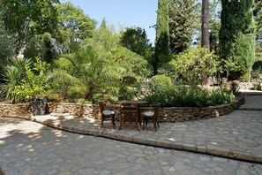 A stone patio with a table and chairs, surrounded by a variety of greenery and trees.