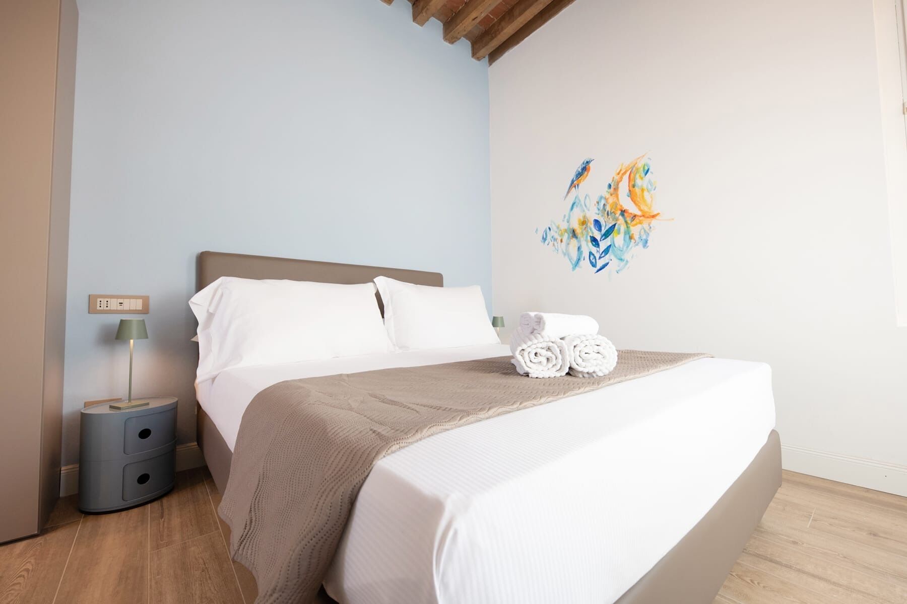 A neatly made bed with a beige blanket, a wall with a colorful abstract painting, and a bedside lamp.