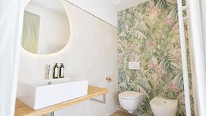 A bathroom with a white sink, two toilets, a mirror, and a wall with a tropical leaf pattern.