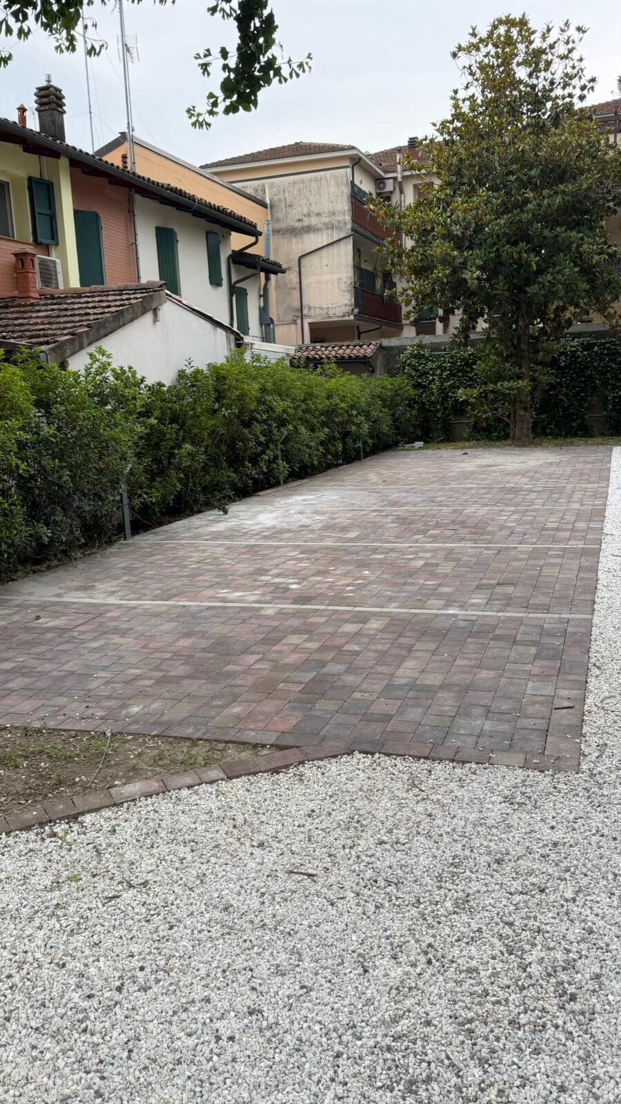 Un patio empedrado con un borde de grava, rodeado de edificios con contraventanas verdes y un árbol.