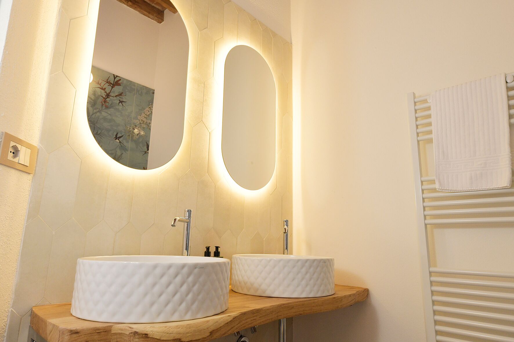 Ein Badezimmer mit zwei runden weißen Waschbecken, einer Holztischplatte, einem großen Spiegel mit sechseckigem Rahmen und einem Handtuchhalter.