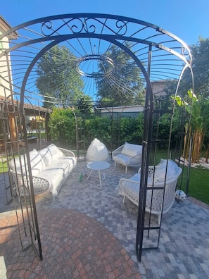 Un área de estar al aire libre techada, con una estructura de pérgola, muebles de mimbre y un patio empedrado.