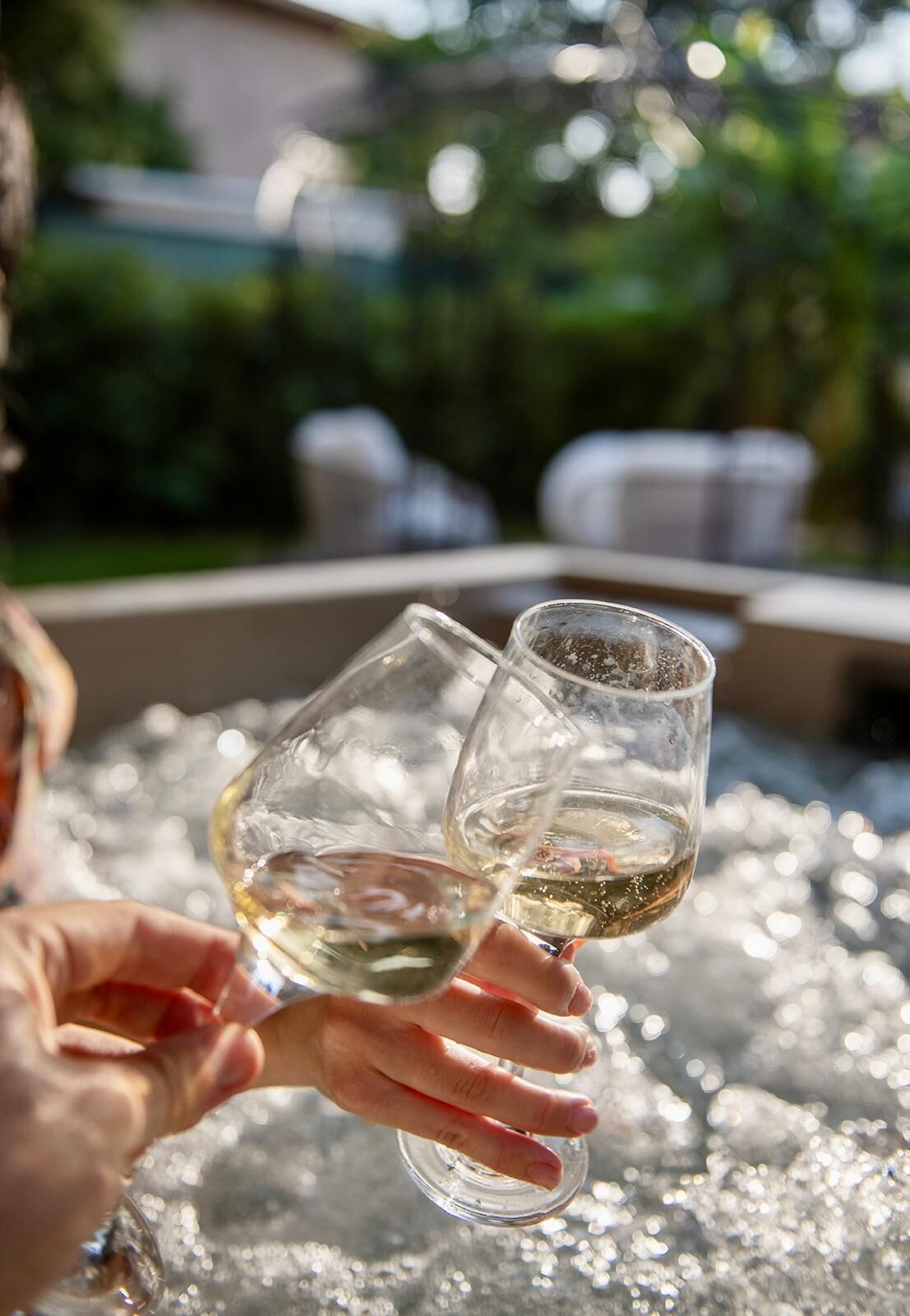 Two people holding wine glasses filled with white wine, with a blurred background of greenery and outdoor seating.