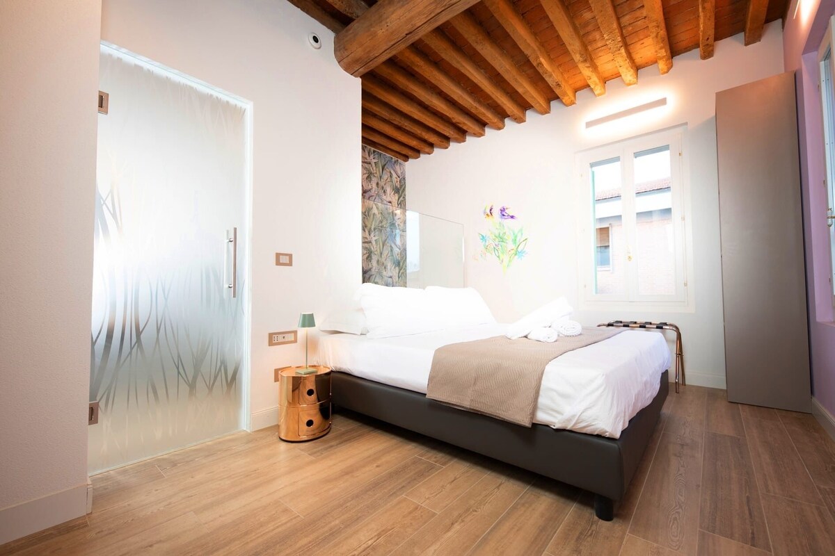 Un dormitorio con cama, suelo de madera, techo con vigas a la vista, pared con un cuadro y ventana con persianas.