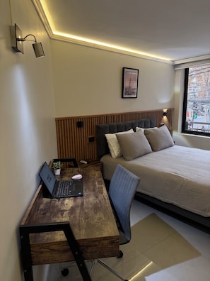 A bedroom with a bed, a desk with a laptop, a chair, a small plant, and a window with a city view. - Exclusivo departamento en Las Condes (Las Condes)