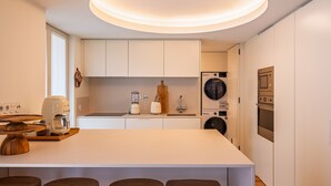 Eine moderne Küche mit weißen Schränken, einer zentralen Kochinsel mit Hockern, einer Kaffeemaschine, einem Mixer, einer Waschmaschine und einem eingebauten Backofen.