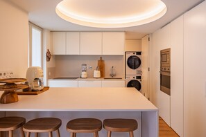 A modern kitchen with white cabinetry, a central island with stools, a coffee machine, a blender, a washing machine, and a built-in oven. - Z - Market Residence 3D (Porto)