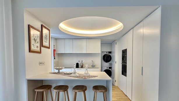 A modern kitchen with white cabinets, a central island with stools, a built-in oven, and a washing machine. - Z - Market Residence 1D (Porto)