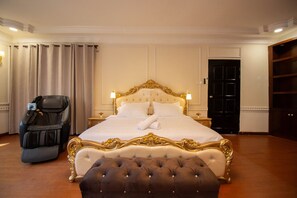 A large bed with ornate headboard, a massage chair, and a black door in a hotel room.