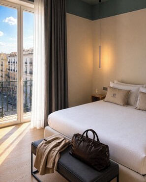 Una habitació d'hotel amb un llit gran, una banca, un bolc, una tauleta de nit i vista a edificis a través d'una finestra.