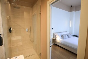A modern bathroom with a glass shower enclosure, a visible toilet, and a bed with white bedding and pillows in the adjacent room. - Altanapoli Rooms & Suites (Napoli)