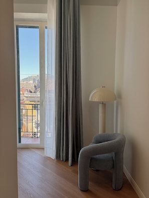 Quarto com vista para a paisagem urbana através de uma janela com cortina, luminária de chão e uma cadeira moderna.