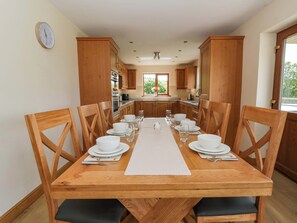 A dining area with a wooden table set for six, surrounded by wooden chairs, in a kitchen with wooden cabinets and a clock on the wall. - Yr Onnen (Rhayader)
