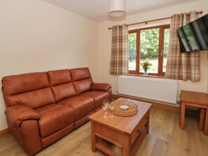A brown leather sofa, a wooden coffee table with glasses and a tray, a small table, and a window with curtains. - Yr Onnen (Rhayader)