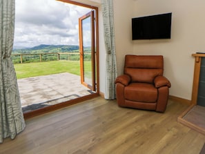 A room with a sliding glass door leading to a patio, a leather armchair, and a mounted flat-screen TV. - Yr Onnen (Rhayader)