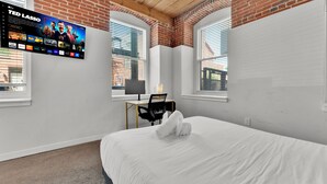 Ein Hotelzimmer mit Bett, Schreibtisch mit Stuhl, Wand-Fernseher und Fenstern mit Jalousien.
