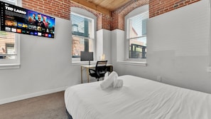 A hotel room with a bed, a desk with a chair, a TV mounted on the wall, and windows with blinds. - Apartmint at N 4th St Philadelphia (Philadelphia)