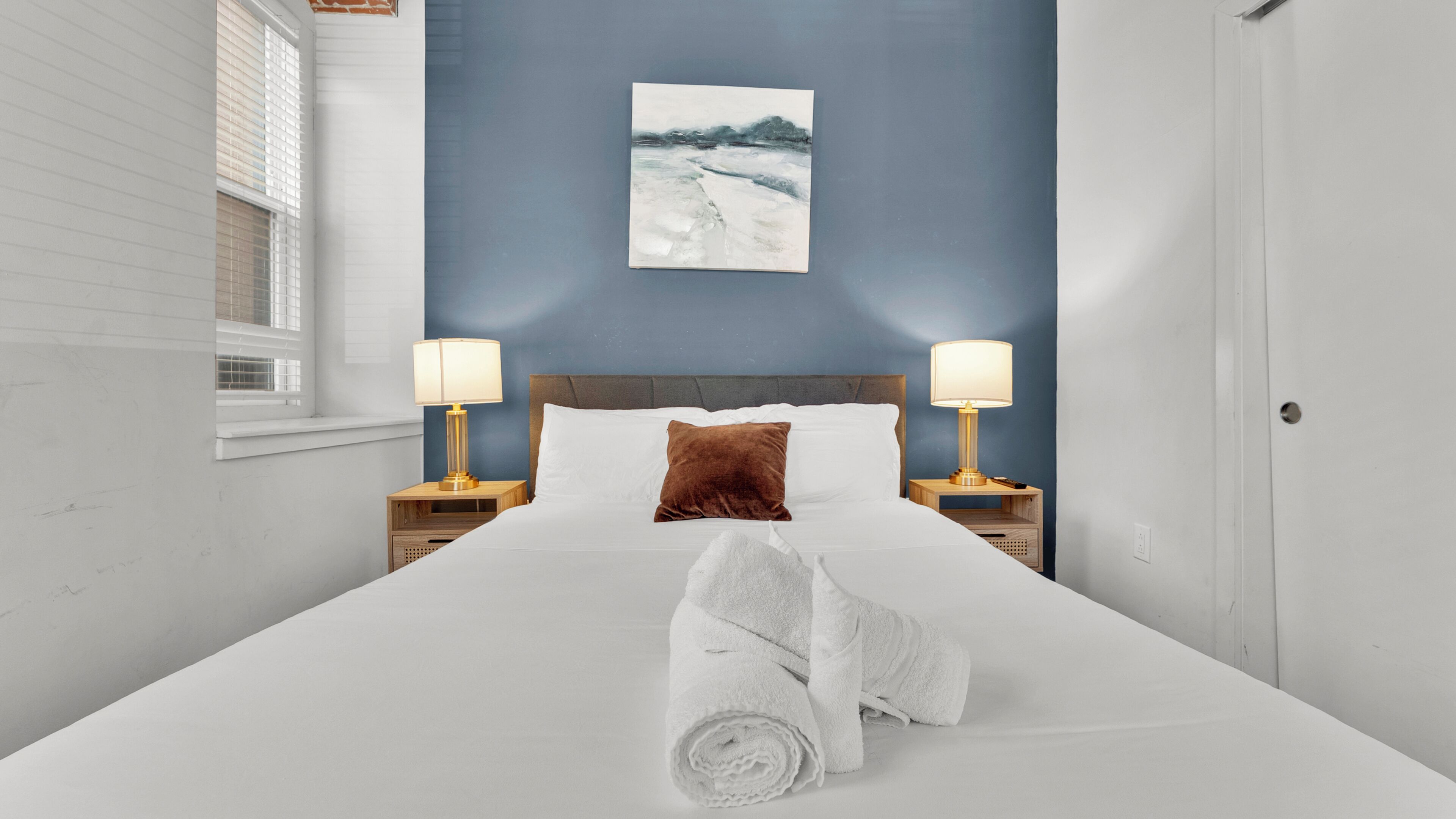 A neatly made bed with a rolled towel, two bedside lamps, a brown pillow, and a painting on the wall.