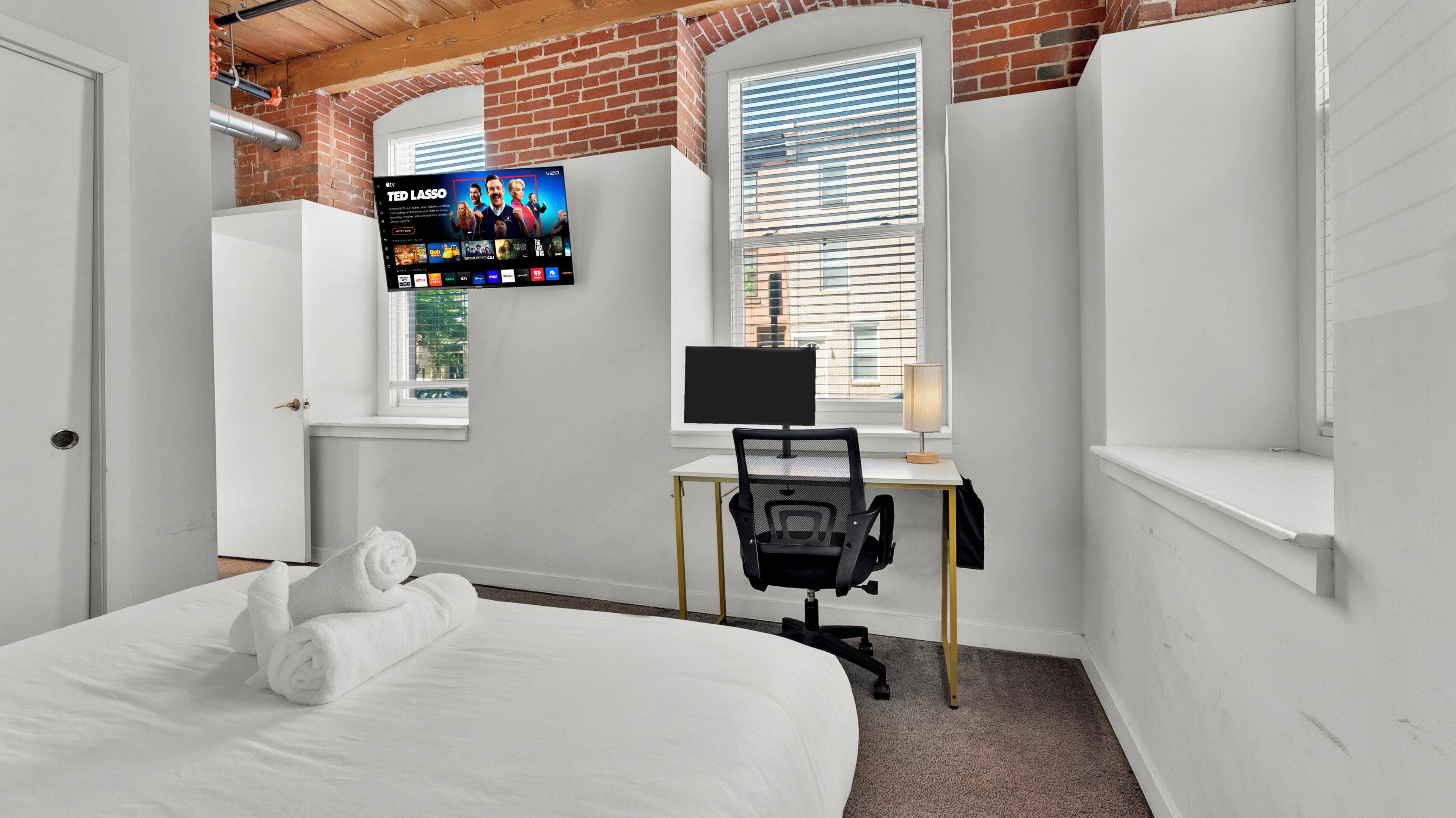 A bedroom with a bed, a desk with a computer, a TV mounted on the wall, and a window with blinds.