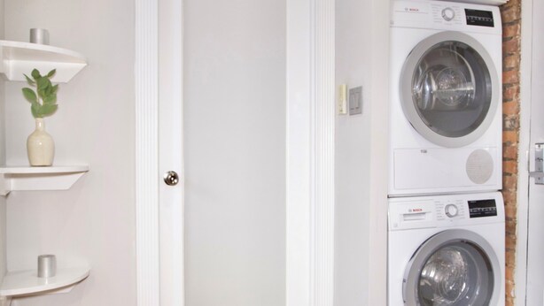 A laundry room with two stacked front-loading washing machines, a white door, and a small shelf with a plant. - New York Style Stay near Central Park (New York)