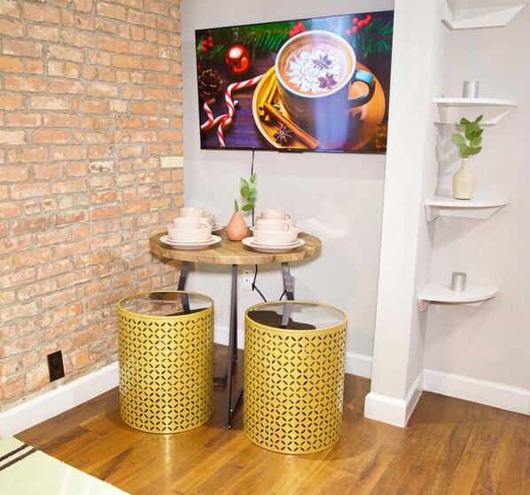 A room with a brick wall, a wooden table with cups and saucers, two yellow patterned trash bins, and a shelf with a plant.