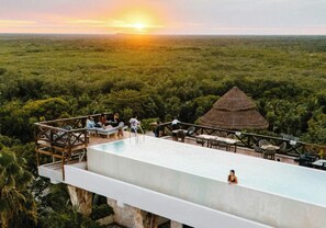 Una piscina infinitum al terrat amb una construcció de teules de palla, envoltada de vegetació abundant i amb vista a un paisatge extens.