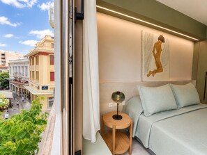 Habitación de hotel con una cama, una mesa redonda de madera, una lámpara de pie, un cuadro en la pared y vista a la calle.