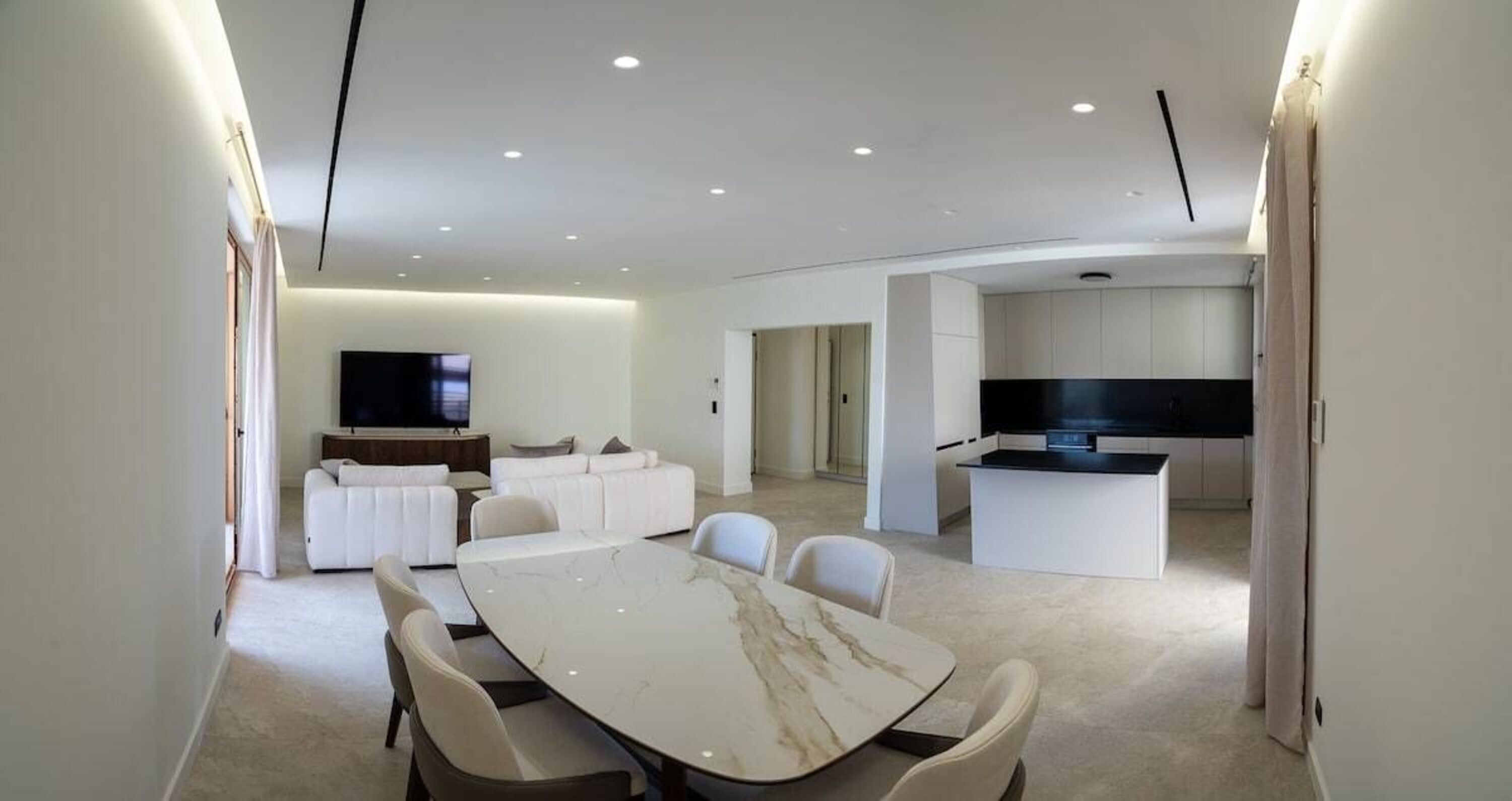 Een moderne woonkamer met een witte zithoek, een eettafel met stoelen, een keuken met een zwart aanrecht en inbouwspots.