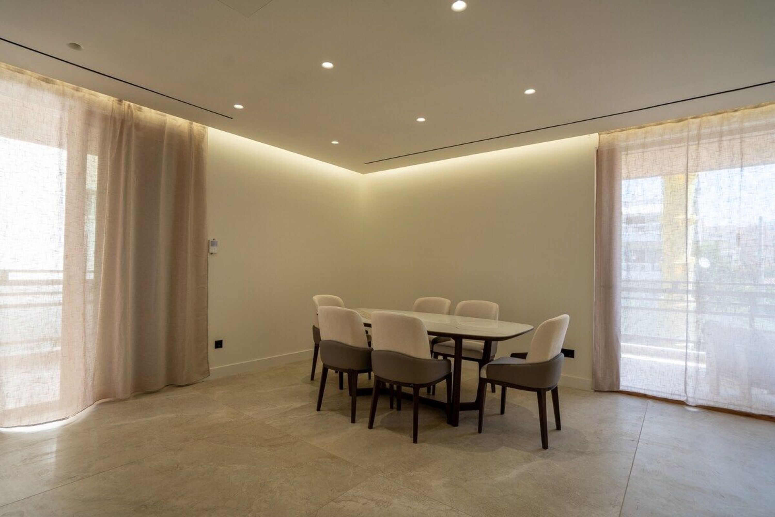 A modern dining area with a round table and six chairs, large windows with sheer curtains, and recessed ceiling lighting.